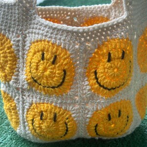 May include: A crocheted tote bag with a white background and yellow smiley faces. The bag has two handles.