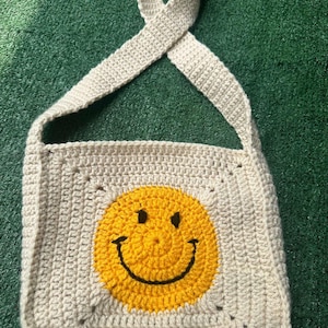 May include: A crocheted beige shoulder bag with a yellow smiley face design.