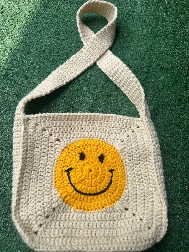 May include: A crocheted beige shoulder bag with a yellow smiley face design.