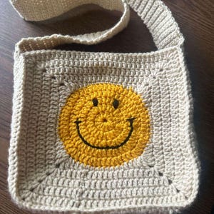 May include: A beige crocheted crossbody bag with a yellow smiley face design.