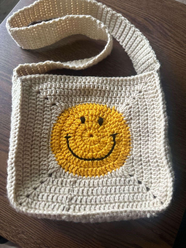 May include: A beige crocheted crossbody bag with a yellow smiley face design.