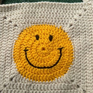 May include: A crocheted square with a yellow smiley face in the center. The square is made with beige yarn and the smiley face is made with yellow yarn.