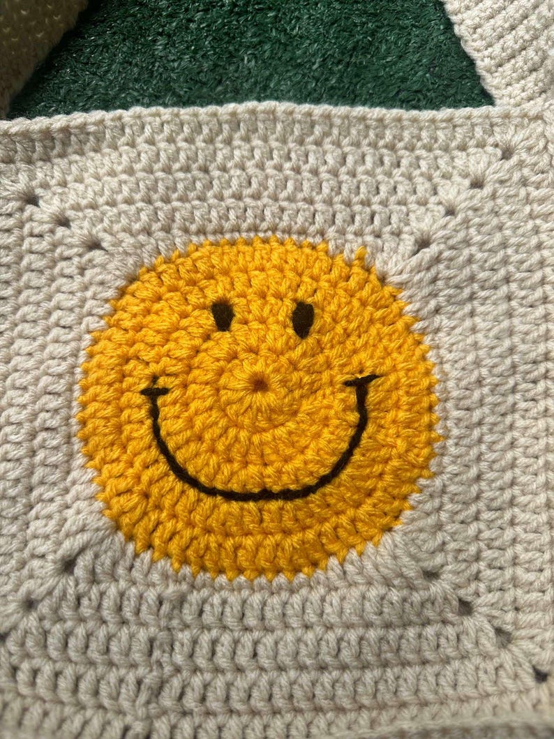 May include: A crocheted square with a yellow smiley face in the center. The square is made with beige yarn and the smiley face is made with yellow yarn.