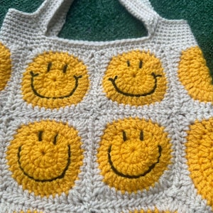 May include: A crocheted tote bag with a white background and yellow smiley faces. The bag has two straps.