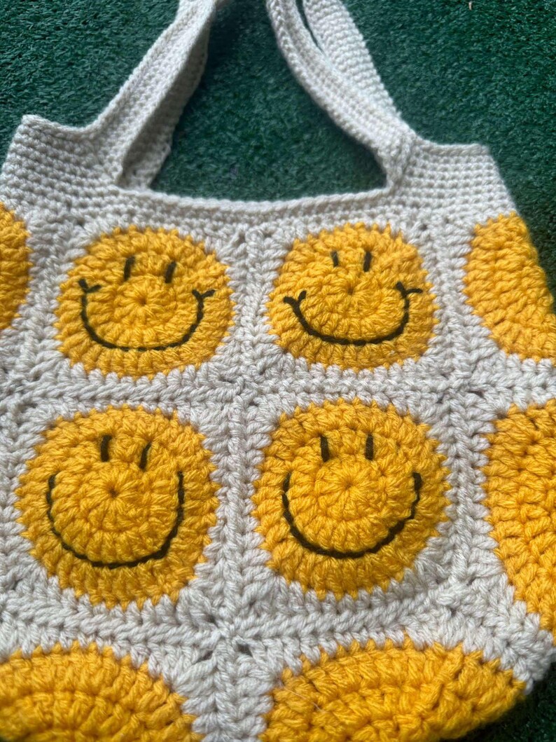 May include: A crocheted tote bag with a white background and yellow smiley faces. The bag has two straps.