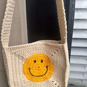 May include: A beige crocheted crossbody bag with a yellow smiley face design.