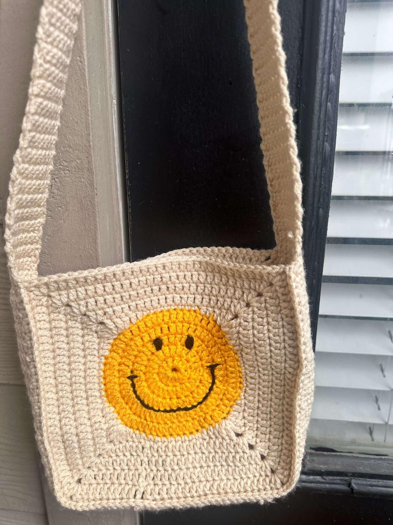May include: A beige crocheted crossbody bag with a yellow smiley face design.