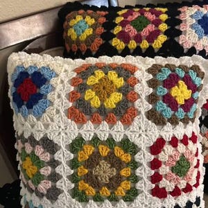 May include: Crocheted pillows with a granny square design. The pillows feature a white background with colorful squares in shades of red, blue, yellow, orange, and green. One pillow has a black border.