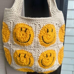 May include: A crocheted tote bag with a beige background and yellow smiley faces. The bag has two straps and is made with a chunky yarn.