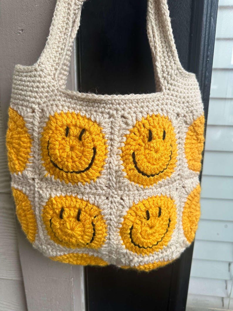 May include: A crocheted tote bag with a beige background and yellow smiley faces. The bag has two straps and is made with a chunky yarn.