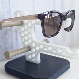 May include: Sunglasses displayed on a white and black 3D-printed stand. The sunglasses have a brown and black tortoiseshell frame and brown lenses. The stand has a honeycomb design and two beige cylindrical arms to hold the glasses.