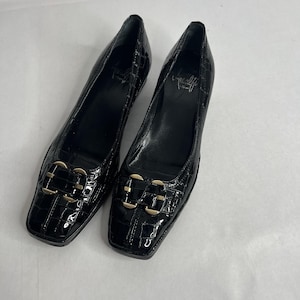 May include: A pair of black, square-toe shoes with a glossy, crocodile-like texture. Each shoe features a gold-colored buckle on the vamp. The shoes are displayed on a white surface.