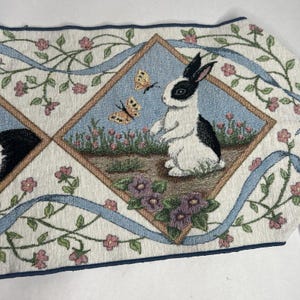 May include: A woven fabric table runner featuring a repeating diamond pattern. Each diamond contains a black and white rabbit standing in a garden with yellow butterflies, surrounded by a border of light blue ribbons and pink floral vines.