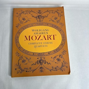 May include: A vintage book cover featuring the text "WOLFGANG AMADEUS MOZART COMPLETE STRING QUARTETS" in a gold oval against a brown background. The cover has ornate black floral and face designs.