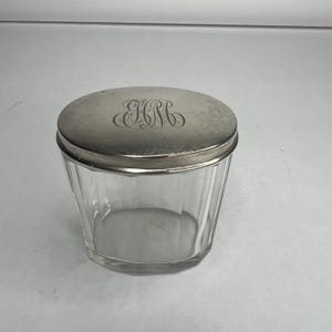 May include: An oval glass jar with vertical fluted sides and a polished silver-toned metal lid. The lid features an intricate, engraved monogram with the letters EHM in a decorative script style, centered on the smooth, metallic surface.