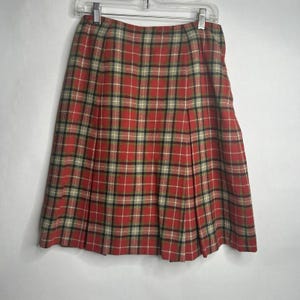 May include: A knee-length pleated skirt featuring a classic red, black, and cream plaid tartan pattern. The skirt is suspended from a silver metal clothing hanger and displays a structured, flared silhouette with vertical pleats throughout the fabric.