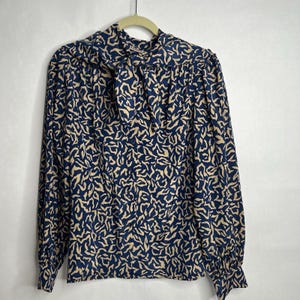 May include: A navy blue blouse with a beige abstract pattern. The long-sleeved top features a bow at the neckline and is hanging on a hanger. The blouse has a vintage style.