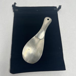 May include: A silver-toned metal shoehorn with a smooth, curved surface and a small circular hole at the top of the handle. The handle features an engraved monogram design. The shoehorn is positioned on a black velvet drawstring pouch.