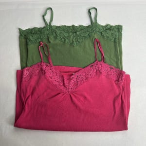 May include: Two camisole tops. One is a vibrant fuchsia pink, the other is a muted green. Both have delicate lace trim along the neckline and thin spaghetti straps. The tops are made of a ribbed knit fabric.