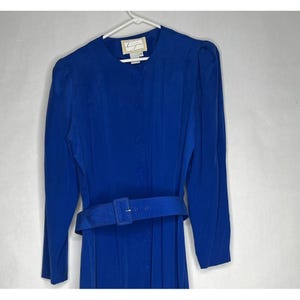 May include: A bright blue dress with long sleeves and a matching belt. The dress has a button-down front and a textured fabric. The belt has a rectangular buckle. The dress is hanging on a white hanger.