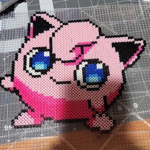 May include: A pink and black pixel art creation of a cartoon character with blue eyes. The character is made of small plastic beads.