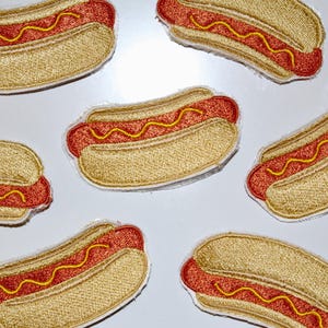 May include: Embroidered patches featuring a hot dog design. The patches have a tan bun, a red hot dog, and yellow mustard. The patches are a fun, food-themed item.