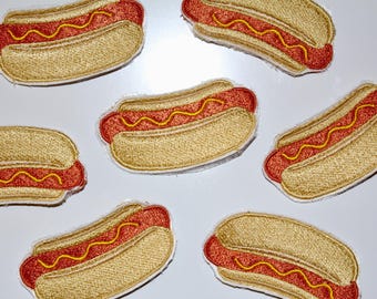 May include: Embroidered patches featuring a hot dog design. The patches have a tan bun, a red hot dog, and yellow mustard. The patches are a fun, food-themed item.