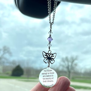 May include: A silver car charm with a butterfly design and a quote from Eleanor Roosevelt. The charm hangs from a chain with purple and clear crystal beads. The quote reads: "The future belongs to those who believe in the beauty of their dreams."