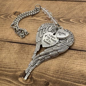 May include: A silver-colored memorial ornament featuring angel wings and heart-shaped charms. One charm reads "a piece of my heart is in heaven," and another says "Mom." The ornament includes a chain and clear beads.