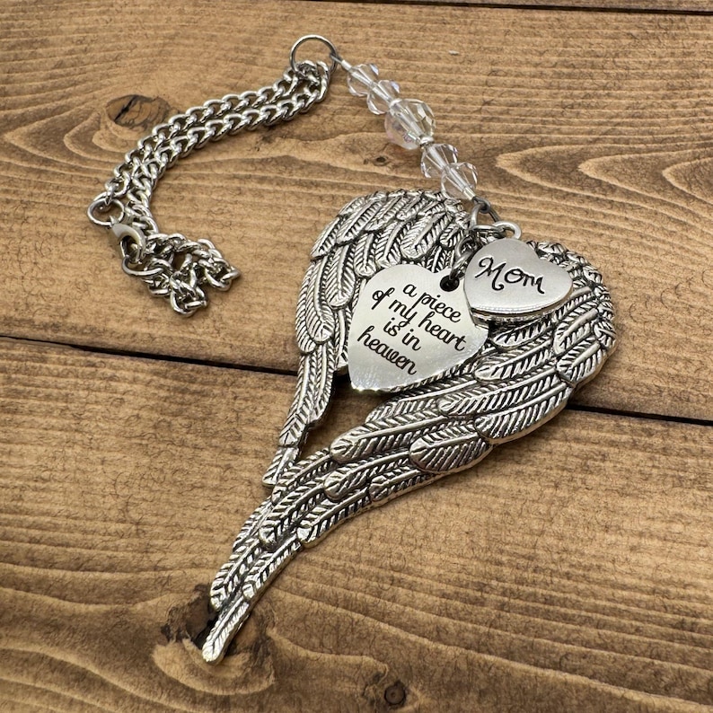 May include: A silver-colored memorial ornament featuring angel wings and heart-shaped charms. One charm reads "a piece of my heart is in heaven," and another says "Mom." The ornament includes a chain and clear beads.