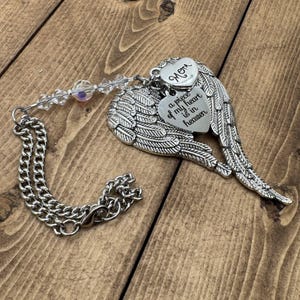 May include: A silver-colored memorial ornament featuring angel wings and two heart-shaped charms. One charm reads "Mom," and the other says "a piece of my heart is in heaven." The ornament includes a chain and a strand of clear beads.