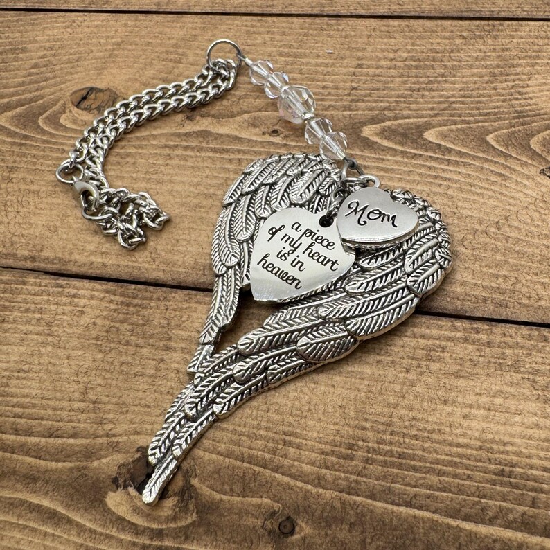 May include: A silver-colored memorial ornament featuring angel wings and heart-shaped charms. One charm reads "Mom," and the other says "a piece of my heart is in heaven." The ornament includes a chain and clear beads.