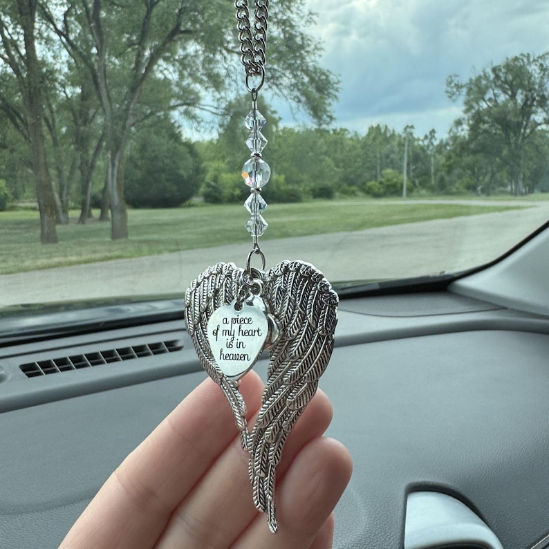 May include: A silver-colored car charm featuring a heart-shaped pendant with angel wings. The pendant has the text "a piece of my heart is in heaven". The charm hangs from a chain with clear crystal beads.