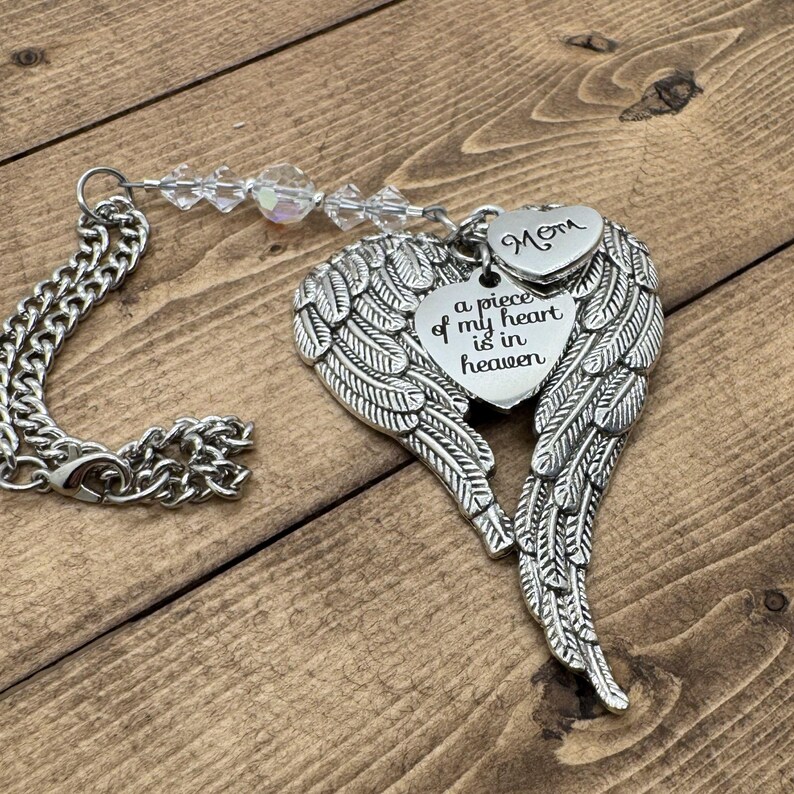 May include: A silver-colored memorial ornament featuring angel wings and a heart-shaped charm. The heart charm is inscribed with the words "Mom" and "a piece of my heart is in heaven." The ornament includes a chain and crystal beads.