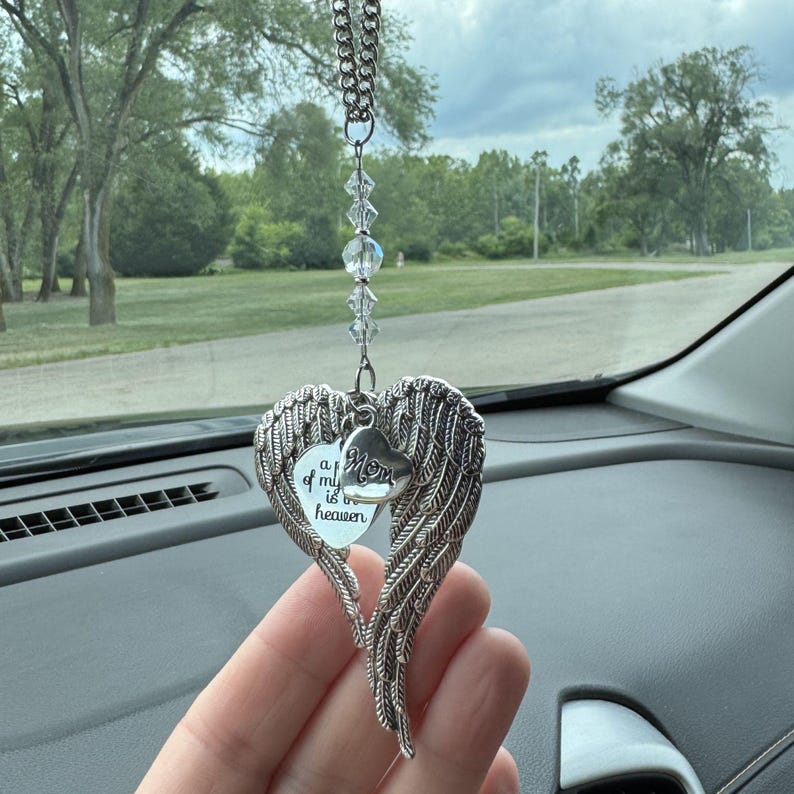 May include: A silver-colored car charm with a heart-shaped pendant featuring angel wings. The pendant has a smaller heart with the word "Mom" and a heart-shaped tag with the text "a of my is in heaven". The charm hangs from a chain with clear beads.