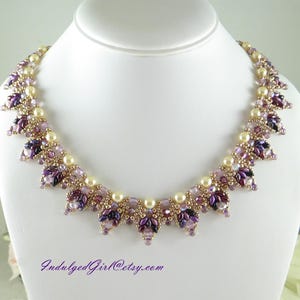 May include: A beaded necklace featuring cream-colored pearls, metallic gold seed beads, and faceted crystals in shades of purple and dark violet. The design incorporates a repeating leaf-like pattern with dangling elements. Text reads IndulgedGirl@etsy.com.
