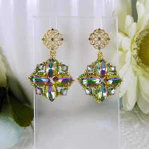 May include: A pair of gold-toned, ornate earrings. Each earring features a filigree top with a small green gem, connected to a square-shaped design with iridescent, multi-colored crystals and green beads. The earrings are displayed on a clear stand.