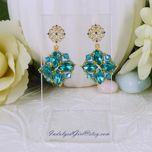 May include: A pair of dangling earrings featuring gold-tone filigree tops and diamond-shaped drops composed of teal crystals and small gold beads. The clear display stand features the text IndulgedGirl@etsy.com printed in blue script at the bottom.
