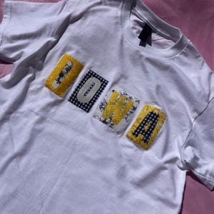 May include: White t-shirt with the word "IOWA" appliquéd across the front. The letters are made from fabric with various patterns, including yellow floral, black and white gingham, and a black and white grid.