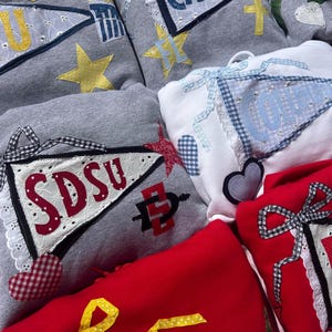 May include: A collection of sweatshirts in various colors, including gray, white, and red. Each sweatshirt features decorative pennant-style patches with text and star or heart accents. Some patches have gingham or lace trim.