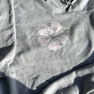 May include: A gray hoodie with a light pink hibiscus flower applique on the front. The flower has a detailed design and is centered on the chest. The hoodie has a drawstring hood and long sleeves.