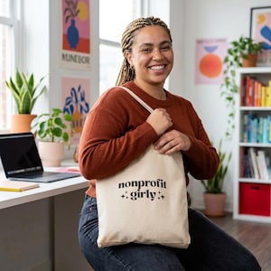 Peut inclure: Un sac fourre-tout beige avec l'inscription "nonprofit girly" en noir. Le sac est tenu par une personne portant un pull de couleur rouille et un jean bleu. L'arrière-plan comprend un ordinateur portable, des plantes et des affiches colorées.
