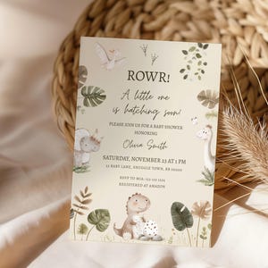 May include: A baby shower invitation with a dinosaur theme. The invitation features illustrations of dinosaurs, foliage, and the text "ROWR! A little one is hatching soon!" The invitation is for Olivia Smith's baby shower, scheduled for November 23rd.