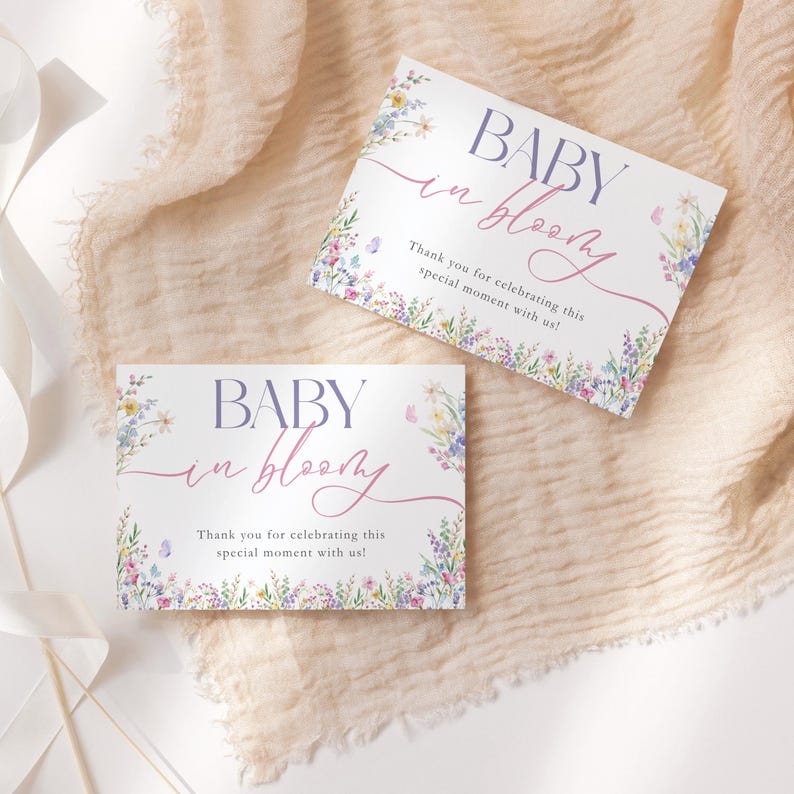 May include: Two white cards with the text "BABY in bloom" in purple and pink script, surrounded by a floral border. The cards also include the text "Thank you for celebrating this special moment with us!"