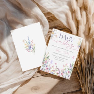 May include: Two white cards with floral designs. One card features a watercolor bouquet, the other reads "BABY in bloom" with event details. The invitation is for a baby shower honoring Olivia Faith.