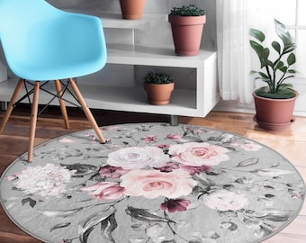 Puede incluir: Una alfombra redonda gris con un diseño floral de acuarela, que incluye rosas rosas y blancas y hojas de color verde oscuro. Una silla azul claro con patas de madera está en el fondo. La alfombra es un elemento decorativo para el hogar.