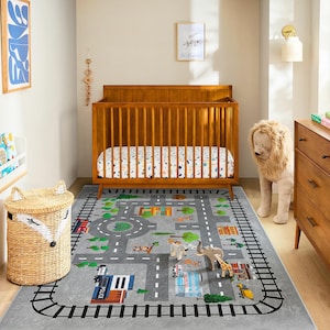 Può includere: Un tappeto grigio a tema stradale con un bordo nero a binario. Il tappeto presenta strade, edifici e alberi. Una culla in legno e un leone di peluche sono sullo sfondo. Un cesto intrecciato con orecchie di volpe è in primo piano.