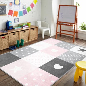 May include: A colorful area rug with a patchwork design featuring squares of pink, gray, and white. The rug has polka dots, hearts, and star patterns. The room also includes a whiteboard, storage baskets, and a yellow stool.