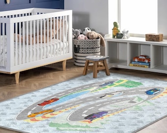 Op de afbeelding: Een lichtblauw kinderkleed met een racebaanontwerp. Het kleed heeft een grijze weg met kleurrijke raceauto's, groen gras en gele en oranje accenten. Het kleed ligt in een kinderkamer.