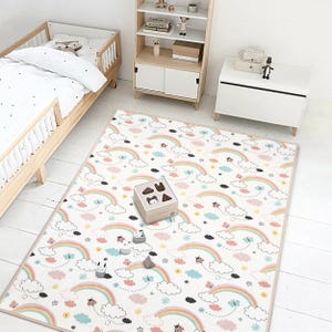 May include: A rectangular play mat with a rainbow and cloud pattern. The mat features pastel rainbows, clouds, butterflies, and ladybugs on a white background. A wooden shape sorter and toy cars are on the mat. A wooden bed and storage furniture are in the background.